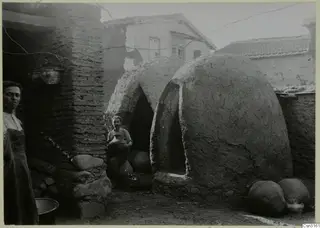 Domed pottery kilns with arched openings in a yard, with a man holding a jar and a woman at the left edge.