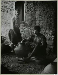 Potter turning a large clay jar outside a stone and mud-brick building, with a second man standing in the doorway behind him.