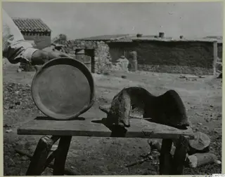 Round fired clay dish and a curved clay windbreak on a wooden bench, with village houses behind at Kliron
