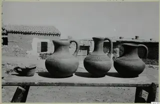 Three jugs and a small handled cup on a rough wooden table, with village houses behind, Kliron