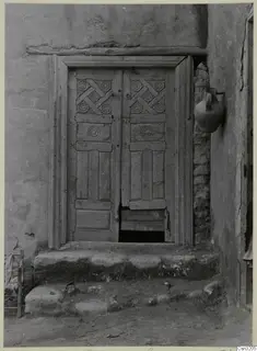 Carved wooden double door in a plastered wall, with worn stone steps below and a clay pot hanging to the right