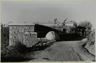 Stone house and a domed whitewashed structure beside a dirt road, with trees behind, in Cyprus