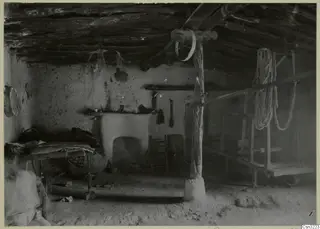 Interior of a plastered room with a log ceiling, a central wooden post, a low frame with bedding and a wooden frame on the right.