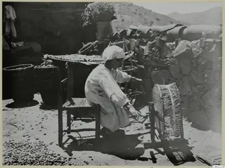 Person in a headscarf and striped smock working at a wooden frame in a yard beside a stone wall.