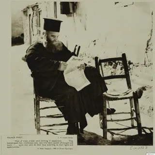 Village priest in a dark robe and tall black hat, seated on a wooden chair and reading a printed sheet beside a whitewashed wall.