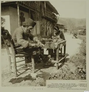 Two men working at a wooden table outside a building, one seated on a chair with pieces of leather on the ground