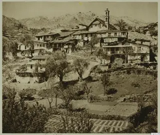 Mountain village of plastered houses with tiled roofs and a tall bell tower, above terraced fields