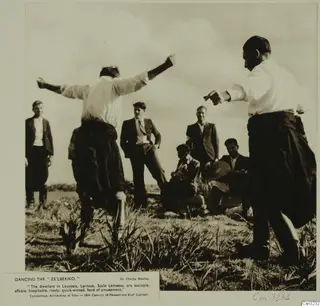 Men dancing in a grassy field while others watch, shown as a photograph mounted on a printed page with a caption.