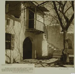 House in Athienou with an arched doorway, a wooden balcony and an outside flight of stone steps, beside a bare tree