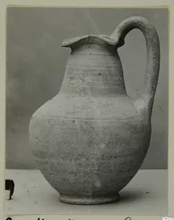 Plain pale pottery jug with a strap handle and flared, wavy rim, photographed on a light surface