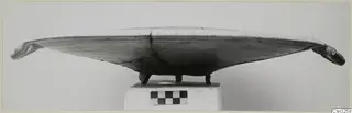Shallow pottery vessel with upturned knobbed ends, standing on three small feet, photographed side-on on a plinth.