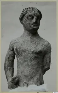 Terracotta figurine of a standing male figure with one arm broken off, shown against a plain grey background