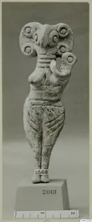 Standing terracotta figurine with a wide head decorated with round discs and one arm raised, shown on a block base