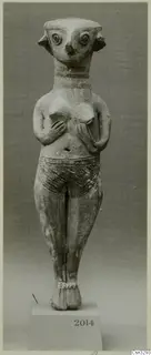 Standing terracotta figurine with large round eyes, a beak-like nose, hands on its chest and an incised belt