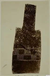 Flat idol-shaped object with incised herringbone and hatched band decoration on a pale background.