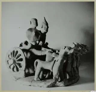 Small terracotta model of two standing figures on a wheeled frame, with a spoked wheel at left and animals in front.