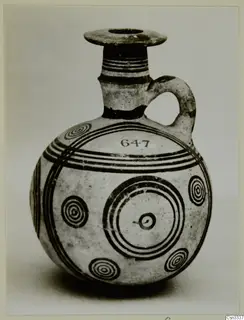 Round pottery juglet with a flared rim, one handle and dark concentric circles and bands painted on a pale body.