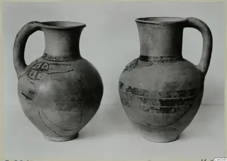 Two handled pottery jugs with dark painted bands and motifs, photographed side by side against a grey background.