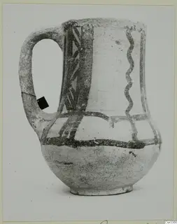 Wheel-made painted jug with a strap handle and dark painted bands, zigzag and vertical pattern on a pale body.