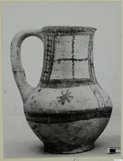 Painted pottery jug with a strap handle, dark bands and panels of dots and cross lines on a pale body.