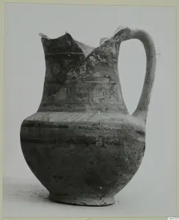 Bichrome wheel-made jug with a strap handle and a broken, uneven rim, photographed against a plain background.