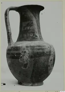Painted pottery jug with a strap handle and dark banded decoration, photographed as a find against a grey backdrop.