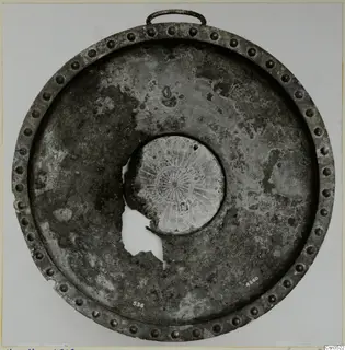 Round metal object with a studded rim, a loop handle and an engraved central boss