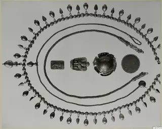 Gold jewellery laid out on a pale background: a bead and pendant necklace, two chains, a relief plaque, a broken bowl and a mesh disc