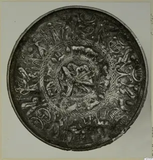 Round metal bowl with raised figures, animal shapes and twisted bands in concentric zones, photographed on a pale card.