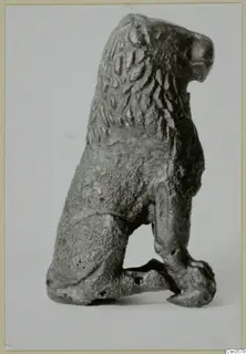 Small bronze figure of a seated animal with a textured mane, shown in side view on a pale backdrop