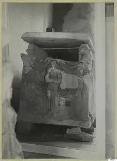 Stone sarcophagus with a carved standing figure on its front, lid slid askew, photographed indoors against a plain wall