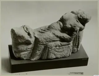 Carved limestone sculpture of a reclining draped figure with head resting on one arm, on a dark stand