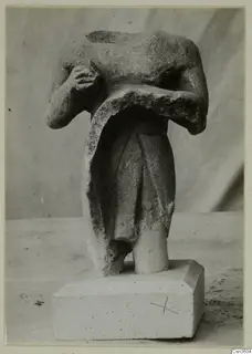 Headless limestone figurine of a standing figure with arms across the chest, on a rectangular base.