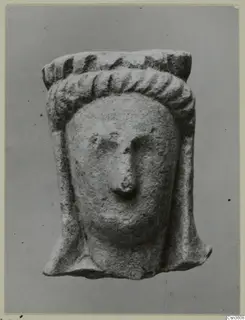 Limestone figurine of a head with ridged headdress and long side locks, shown against a plain grey backdrop.