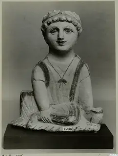 Seated limestone figurine with curly hair, painted eyes and dark painted straps, shown on a dark wooden base.