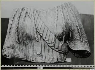 Marble sculpture fragment of a draped garment with vertical pleats and a diagonal band, shown on a dark stand.