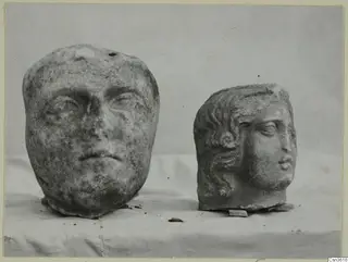 Two carved marble heads side by side on a pale cloth, one broad-faced and one smaller with curled hair