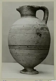 Pottery jug with a single handle, a tall neck and horizontal dark bands on a pale body, on a stand.