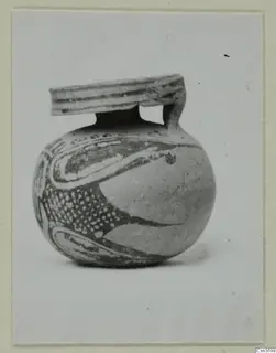 Small round painted pottery vessel with a flat handle arching over its mouth, shown against a light background