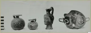 Four painted pottery vessels in a row on a cloth, including a juglet with a modelled head and a broken two-handled cup
