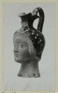 Jug shaped as a female head with a strap handle and trefoil mouth, shown on a mounted card