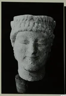 Limestone head of a sculpture from Mersinaki, with carved curls across the top and a worn face, set on a wooden support.