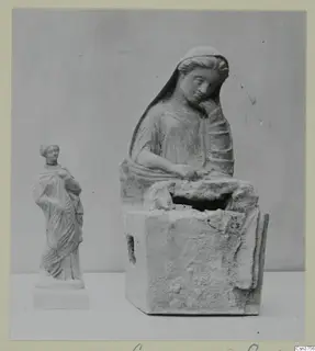 Two pale female figures photographed together: a larger seated figure on a block base and a small standing draped figure.