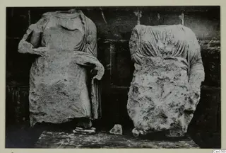 Two headless stone statues of draped standing figures against a dark wall, from Arsos