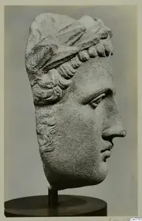 Carved stone head of a figure in profile facing right, with layered hair bands, on a dark stand.