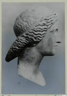 Stone head in profile facing right, with carved wavy hair bands and a long roll of hair falling behind the ear