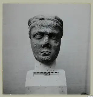 Carved stone head from a statue, shown from the front on a white pedestal with a scale bar