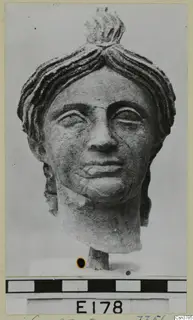 Carved stone head of a woman with hair swept up in a topknot, on a short base against a grey backdrop.