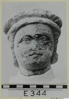 Stone head sculpture seen from the front, with a thick rolled headdress, large eyes and a pitted, worn surface