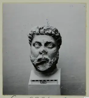 Roman sculpture of a bearded man's head with curly hair, set on a square pedestal.